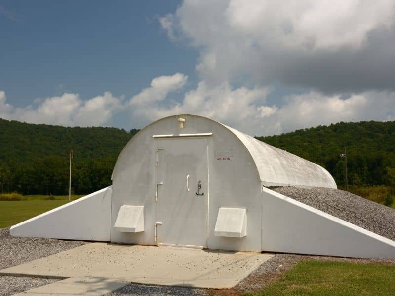 Home survey detached storm shelter ringgold | Ringgold Land Surveying White dome-style shelter with a metal door, flanked by low concrete ramps, set on a gravel pad in a grassy, hilly landscape.